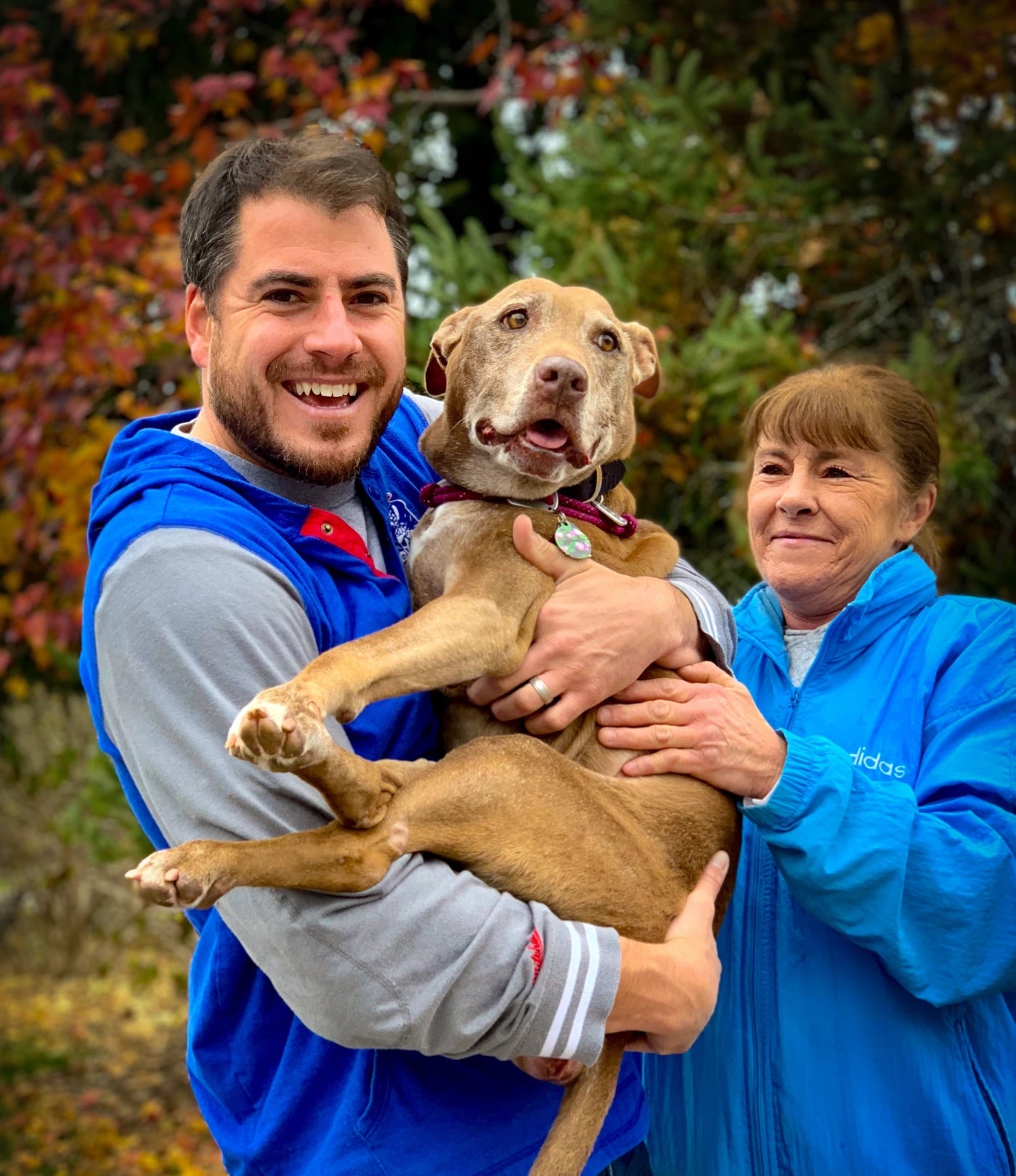 two people standing wearing blue jackets, smiling, holding medium size dog in their arms.